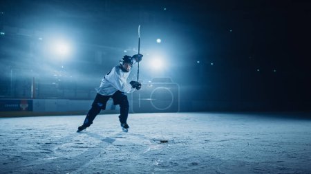 Foto de Ice Hockey Rink Arena: Jugador profesional Disparos, golpes, Stricking the Puck con palos de hockey. Atleta anotando una meta. Captura amplia dramática, iluminación cinematográfica. - Imagen libre de derechos