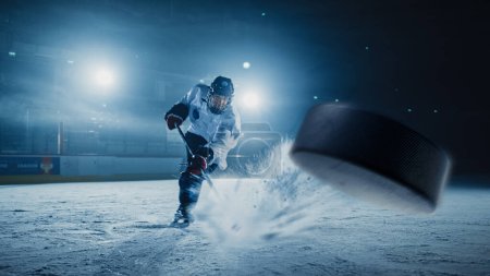 Téléchargez les photos : Patinoire de hockey sur glace : Joueur professionnel tirant sur la rondelle avec bâton de hockey. Focus sur 3D Flying Puck avec effet Flou Motion. Prise de vue large dramatique, éclairage cinématographique. - en image libre de droit