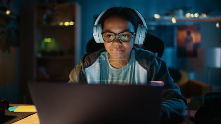 Photo for Young Teenage Multiethnic Girl Using Laptop Computer and Wearing Headphones in a Dark Cozy Room at Home. She's Browsing Educational Research Online. Studying Science School Homework Concept. - Royalty Free Image