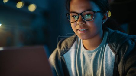 Téléchargez les photos : Jeune adolescente noire multiethnique utilisant un ordinateur portable dans une chambre confortable sombre à la maison. Elle navigue sur Recherche en éducation en ligne. Étude du concept des devoirs de l'École des sciences. - en image libre de droit