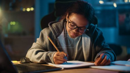 Photo for Young Teenage Multiethnic Black Girl Writing Down Homework in a Notebook with a Pencil, Using Laptop Computer in a Dark Cozy Room at Home. She's Browsing Educational School Research Online. - Royalty Free Image