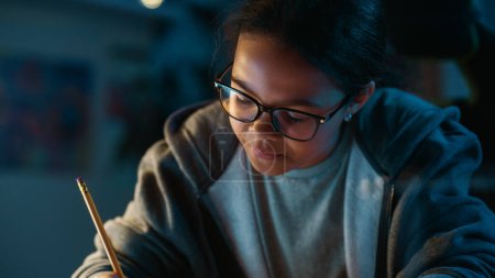 Foto de Joven adolescente multiétnica chica negra escribiendo tareas en un cuaderno con un lápiz, utilizando el ordenador portátil en una habitación oscura y acogedora en casa. Ella está explorando la investigación de la escuela educativa en línea. - Imagen libre de derechos