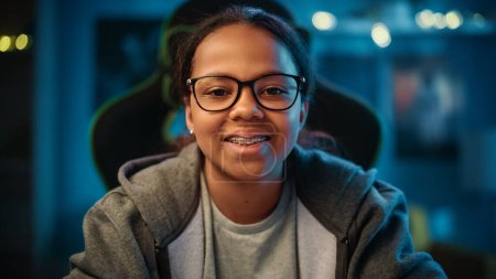 Téléchargez les photos : Portrait d'une jolie jeune adolescente noire multiethnique dans une chambre sombre et confortable à la maison. Elle est heureuse, regarde la caméra et sourit. Jeune femme porte des lunettes et des bretelles. - en image libre de droit