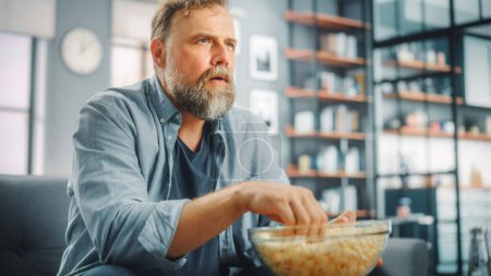 Photo for Handsome Middle Aged Bearded Man Sitting on a Couch Watches TV at Home. Focused Fan Eats Snacks, Watches TV Shows, Sports Event, Film, On Demand Streaming Service. - Royalty Free Image