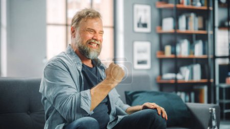 Photo for Charismatic Bearded Man Sitting on a Couch Watches Game on TV, Celebrates Sports when Team Wins Championship. Focused, Joyfully Intense and Authnetic Fan Cheers when Favourite Club Play - Royalty Free Image