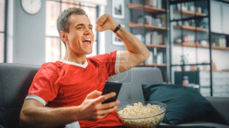Charismatic Young Adult Man Sitting on a Couch Watches Game on TV, uses Smartphone App for Score, Bet, Statistics, Celebrates Victory when Team Wins Championship. Happy Fan Watches Sport.