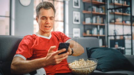 Charismatic Young Adult Man Sitting on a Couch Watches Game on TV, uses Smartphone App for Score, Bet, Statistics, Celebrates Victory when Team Wins Championship. Happy Fan Watches Sport.