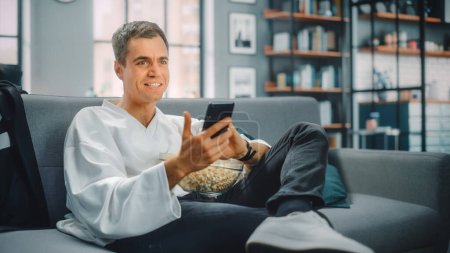 Handsome Man Sitting on a Couch Watches Ice Hockey Game on TV, uses Smartphone App for Score, Bet, Statistics, Eats Snacks, Watching Championship. Happy Fan Watches Sport.