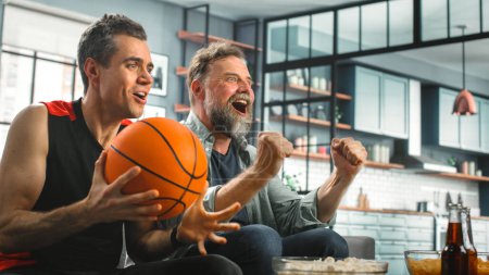 Photo for At Home Two Fans Sitting on a Couch Watch Basketball Game on TV, Celebrate Scoring Victory. Thier Team Wins Championship. Friends Watch Favourite Club Play - Royalty Free Image