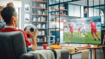 Photo for At Home Soccer Fans Sitting on a Couch Watch Football Game on TV, Cheer for Favourtite Sports Team to Win Championship. Screen Shows Professional Football Club Play. Over the Shoulder - Royalty Free Image