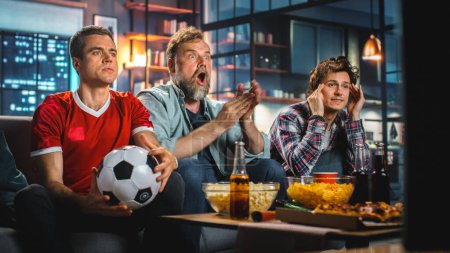 Foto de Night At Home: Three Joyful Soccer Fans Sentado en un juego de reloj de sofá en la televisión, Celebra la victoria cuando el equipo deportivo gane el campeonato. Grupo de Amigos Animo por Club de Fútbol Favorito Jugar. - Imagen libre de derechos
