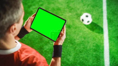 Téléchargez les photos : Lecteur de football professionnel tenant ordinateur tablette numérique avec écran vert Modèle de clé chroma. Haut haut vers le bas au-dessus de prise de vue du contenu de navigation d'étoile de football tandis que debout sur le terrain de jeu - en image libre de droit