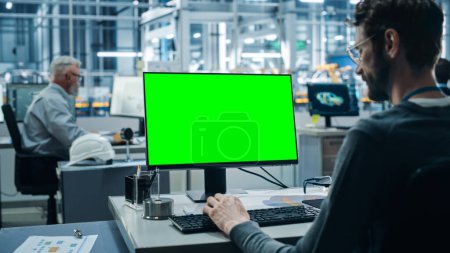 Photo for Car Factory Office: Caucasian Male Automotive Engineer Sitting at His Desk Working on Green Screen Chroma Key Computer. Automated Robot Arm Assembly Line Manufacturing. Over Shoulder Shot - Royalty Free Image