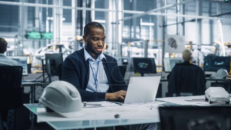 Junger afroamerikanischer Ingenieur arbeitet am Laptop in einem Büro in einem Montagewerk. Industriespezialist arbeitet an Fahrzeugteilen in technologischer Entwicklungseinrichtung.
