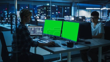 Vielfältige Teamarbeit im Büro bei Nacht: Behinderte arbeiten am Green Screen Chroma Key Computer mit prothetischem Arm. Team von Software-Ingenieuren arbeitet spät an innovativer App