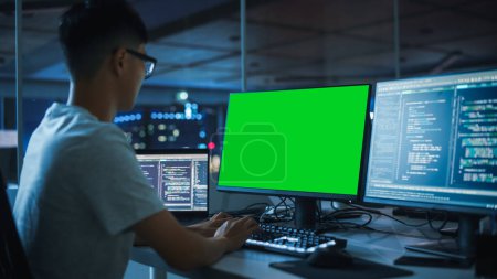 Photo for Night Office: Young Japanese Man in Working on Green Screen Chroma Key Desktop Computer. Team of Programmers Typing Code, Creating Modern Software, e-Commerce App Design, e-Business Programming - Royalty Free Image