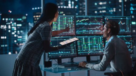 Financial Analyst Talking to Investment Banker in the Evening at Work. Chatting About Real-Time Stock Chart Data on Multi-Monitor Workstation. Businesspeople Have a Meeting in Broker Agency Office.