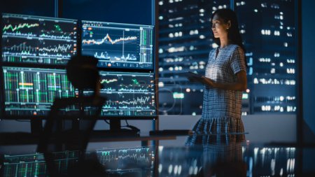 Financial Analyst Using Tablet Computer, Standing Next to Multi-Monitor Workstation with Real-Time Stocks, Commodities and Exchange Market Charts. Businesswoman Working in Investment Bank City Office.