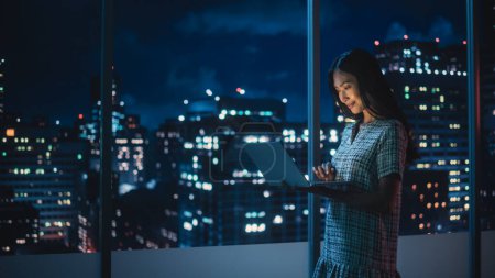 Financial Analyst Using Laptop Computer, Standing Next to Window with Beautiful Night City Skyline with Skyscrapers. Confident Asian Businesswoman Working in Modern Corporate Office.