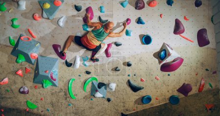 Foto de Escalador experimentado fuerte practicando escalada en solitario en la pared de Bouldering en un gimnasio. Ejercicio del hombre en el gimnasio interior, haciendo deporte extremo para su entrenamiento de estilo de vida saludable. Disparo desde la espalda. - Imagen libre de derechos