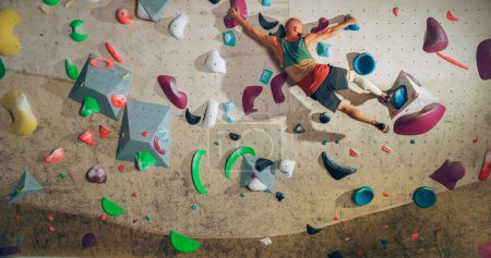 Starker erfahrener Bergsteiger übt sich im Solo-Klettern an der Boulderwand in einer Turnhalle. Der Mann trainiert in einem Indoor-Fitnessstudio und macht Extremsport für sein gesundes Lifestyle-Training. Schuss von hinten.