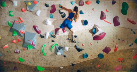 Photo for Athletic Female Rock Climber Practicing Solo Climbing on Bouldering Wall in a Gym. Female Exercising at Indoor Fitness Facility, Doing Extreme Sport for Her Healthy Lifestyle Training. Shot from Back. - Royalty Free Image