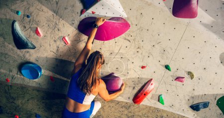 Photo for Athletic Female Rock Climber Practicing Solo Climbing on Bouldering Wall in a Gym. Female Exercising at Indoor Fitness Facility, Doing Extreme Sport for Her Healthy Lifestyle Training. Shot from Back. - Royalty Free Image