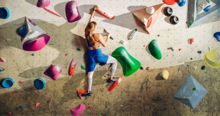 Atlética escaladora femenina practicando escalada en solitario en la pared de roca en un gimnasio. Ejercicio femenino en el gimnasio interior, haciendo deporte extremo para su entrenamiento de estilo de vida saludable. Disparo desde la espalda.