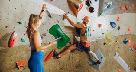 Kletterlehrer gibt einem Anfänger Unterricht an einer Boulderwand in einer Turnhalle. Sportlerin hält Tablet-Computer in der Hand und zeigt starken, fitten Männern korrekte Klettertechniken.