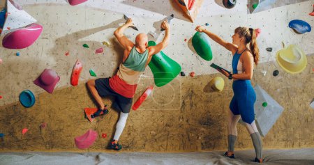 Kletterlehrer gibt einem Anfänger Unterricht an einer Boulderwand in einer Turnhalle. Sportlerin hält Tablet-Computer in der Hand und zeigt starken, fitten Männern korrekte Klettertechniken.