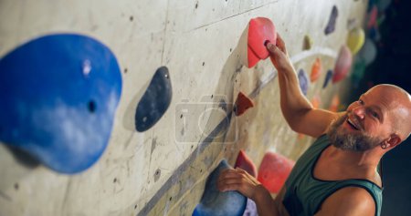Starker erfahrener Kletterer übt sich im Solo-Klettern an der Boulderwand in der Turnhalle. Der Mann trainiert in einem Indoor-Fitnessstudio und macht Extremsport für sein gesundes Lifestyle-Training. Porträt aus nächster Nähe