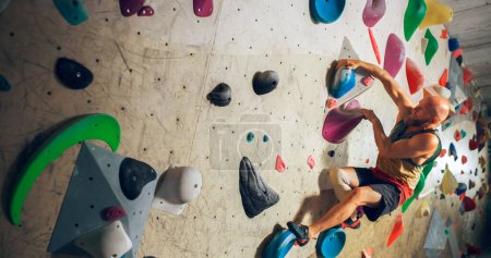 Starker erfahrener Kletterer übt sich im Solo-Klettern an der Boulderwand in der Turnhalle. Mann trainiert im Indoor-Fitnessstudio, macht Extremsport für seinen gesunden Lebensstil.