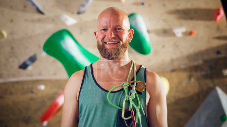 Photo for Strong Masculine Male Athlete Smiling and Posing at Rock Climbing Gym with Bouldering Wall Background. Handsome Happy Bald Man with Ginger Beard, Wearing Colorful Undershirt and Holding Rope Harness. - Royalty Free Image