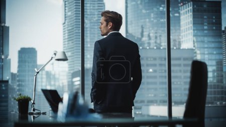 Foto de Pensativo joven hombre de negocios en un traje perfecto a medida de pie en su oficina moderna Mirando por la ventana de la gran ciudad con rascacielos. Estrategia exitosa del proyecto de planificación del gerente de finanzas. - Imagen libre de derechos