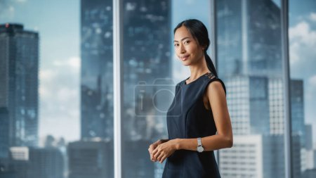 Foto de Hermoso retrato de una mujer de negocios asiática en elegante vestido negro posando junto a la ventana en la oficina de la gran ciudad con rascacielos. Confiada mujer CEO sonriendo. Exitoso Gerente de Negocios Diversos. - Imagen libre de derechos