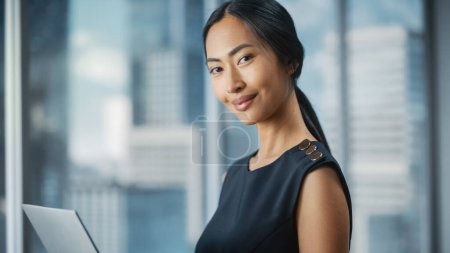 Foto de Hermoso retrato de una mujer de negocios asiática en elegante vestido negro usando computadora portátil, posando junto a la ventana en la oficina de la ciudad. Confiada mujer CEO sonriendo. Exitoso Gerente de Negocios Diversos. - Imagen libre de derechos