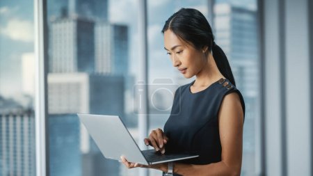 Photo for Beautiful Portrait of an Asian Businesswoman in Stylish Black Dress Using Laptop Computer, Posing Next to Window in City Office. Confident Female CEO Smiling. Successful Diverse Business Manager. - Royalty Free Image