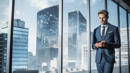 Photo for Confident Young Businessman in a Suit Standing in Modern Office, Using Smartphone, Looking out of the Window on Big City with Skyscrapers. Successful Finance Manager Planning Work Projects. - Royalty Free Image