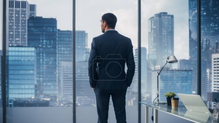 Photo for Thoughtful Young Businessman in a Perfect Tailored Suit Standing in His Modern Office Looking out of the Window on Big City in the Evening. Successful Finance Manager Planning Project Strategy. - Royalty Free Image