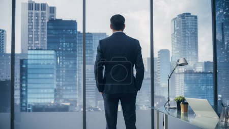 Photo for Thoughtful Young Businessman in a Perfect Tailored Suit Standing in His Modern Office Looking out of the Window on Big City in the Evening. Successful Finance Manager Planning Project Strategy. - Royalty Free Image