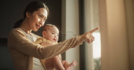 Belle jeune femme asiatique tenant son bébé dans ses bras tout en se tenant à côté d'une fenêtre à la maison. mignon petit bambin reposant dans son mère embrasser