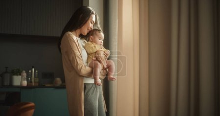 Hermosa mujer asiática joven sosteniendo a su bebé en sus brazos mientras está de pie junto a una ventana en casa. Lindo niño pequeño descansando en su abrazo de las madres