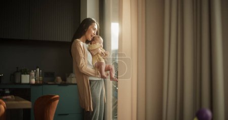 Die schöne junge Asiatin hält ihr Baby im Arm, während sie zu Hause neben einem Fenster steht. Niedliches kleines Kleinkind ruht in der Umarmung seiner Mütter