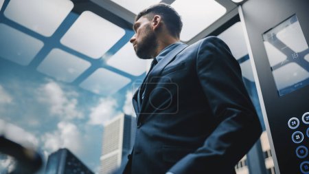 Téléchargez les photos : Homme d'affaires prospère en costume, regardant par la fenêtre, tout en chevauchant ascenseur en verre au bureau dans le centre d'affaires moderne. Beau Top Manager - en image libre de droit