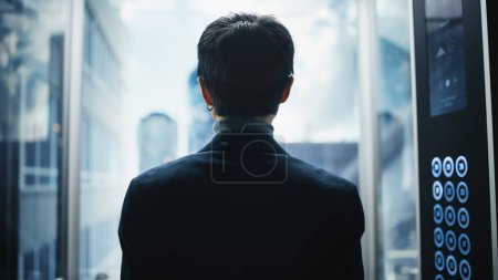 Téléchargez les photos : Ascenseur en verre d'équitation d'homme d'affaires japonais élégant au bureau dans le centre d'affaires moderne. Homme regardant les gratte-ciel du centre-ville moderne - en image libre de droit