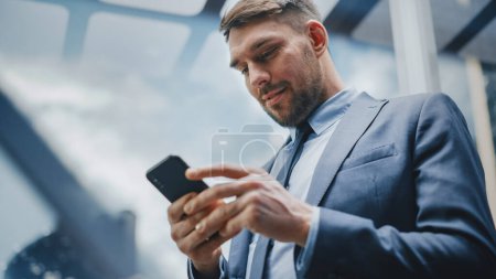 Successful Businessman Riding Glass Elevator to Office in Modern Business Center. Handsome Happy Man Smile while Using Smartphone, Write Text Message