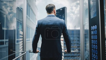 Téléchargez les photos : Homme d'affaires prospère dans un ascenseur en verre d'équitation costume au bureau dans le centre d'affaires moderne. Jeune homme regardant les gratte-ciel modernes du centre-ville - en image libre de droit