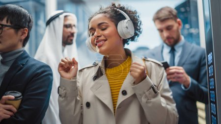 Carefree Black Female Riding Glass Elevator to Office in Modern Business Center. Beautiful Latin Woman is Happy, Smile, Listen to Music on Headphones