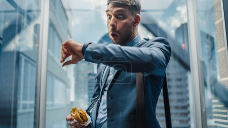 Photo for Business Manager is Late for Work, Riding Glass Elevator in Modern Office Building. Handsome Man Eating a Tasty Take Away Hamburger in a Hurry on the - Royalty Free Image