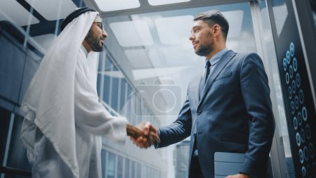 Téléchargez les photos : Homme d'affaires Parler avec un partenaire d'investissement arabe tout en chevauchant ascenseur en verre au bureau dans un centre d'affaires moderne. International Corporate Associates - en image libre de droit